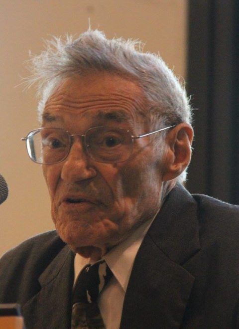 Elderly man in a suit and glasses speaking at a microphone indoors