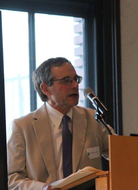 Man speaking at a podium with a microphone, holding papers in a conference room beside a window