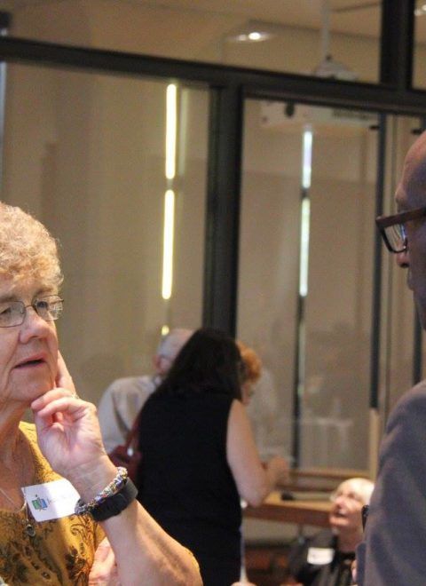 People conversing in a conference room, with glass doors and seated attendees in the background
