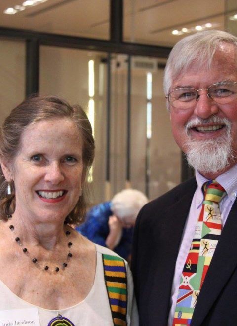 Two smiling people posing indoors, one in a dark suit and colorful tie, the other in a white top and striped strap.