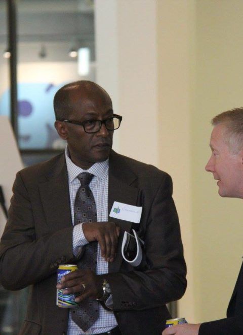 Two men in suits talking in a hallway, one holding a water bottle and wearing glasses.