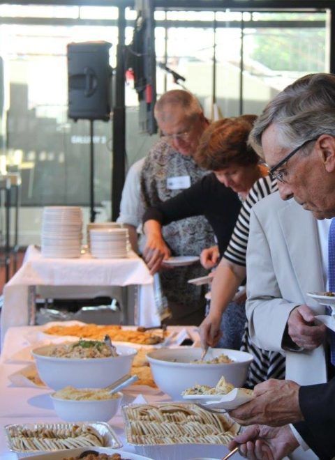 People serving food at a buffet table with bowls and trays in a bright indoor setting