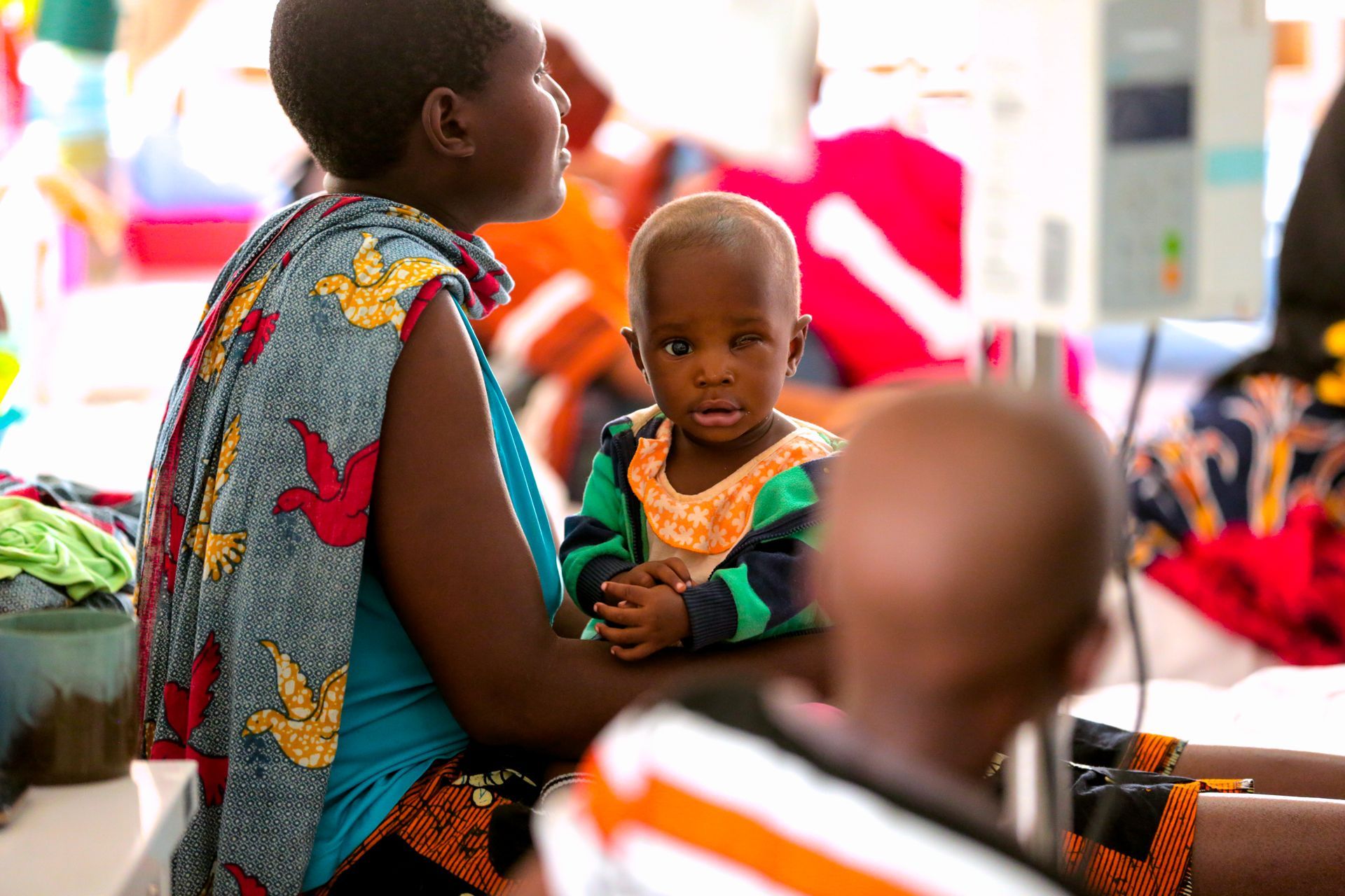 Woman holding child at a colorful market stall, with baskets and blurred shoppers in the foreground