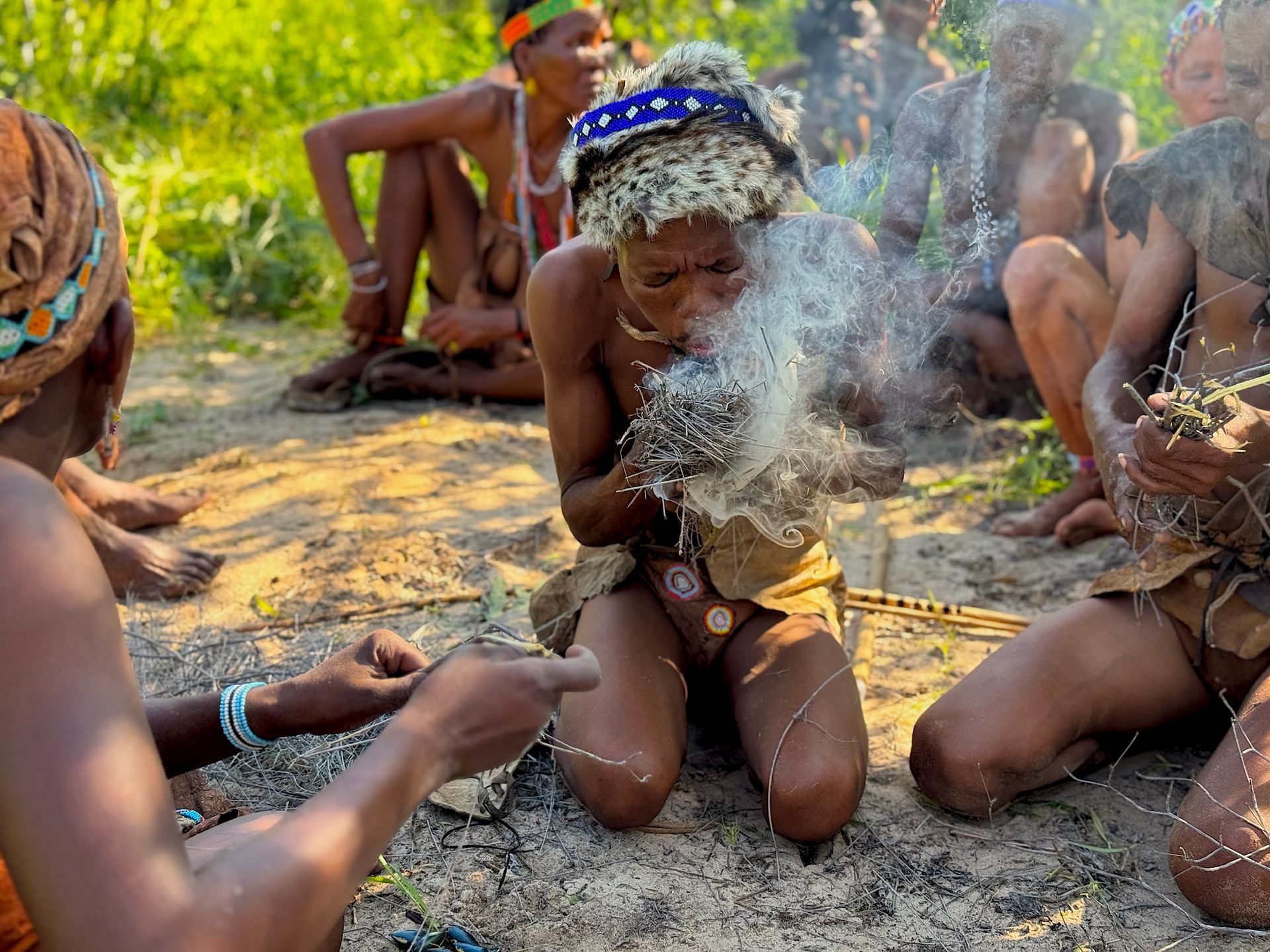 People gathered outdoors around a smoking fire, kneeling and holding bowls in a sandy area.