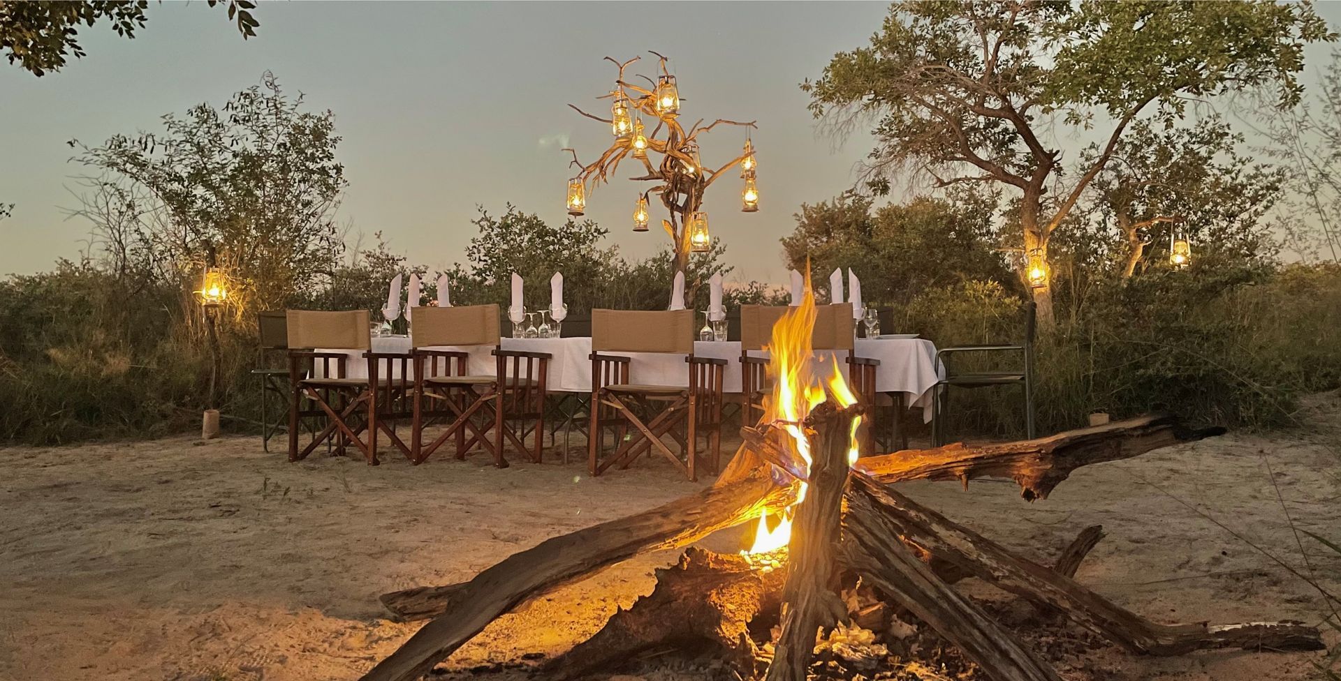 Outdoor dining setup beside a campfire at dusk, with chairs and lanterns under trees.
