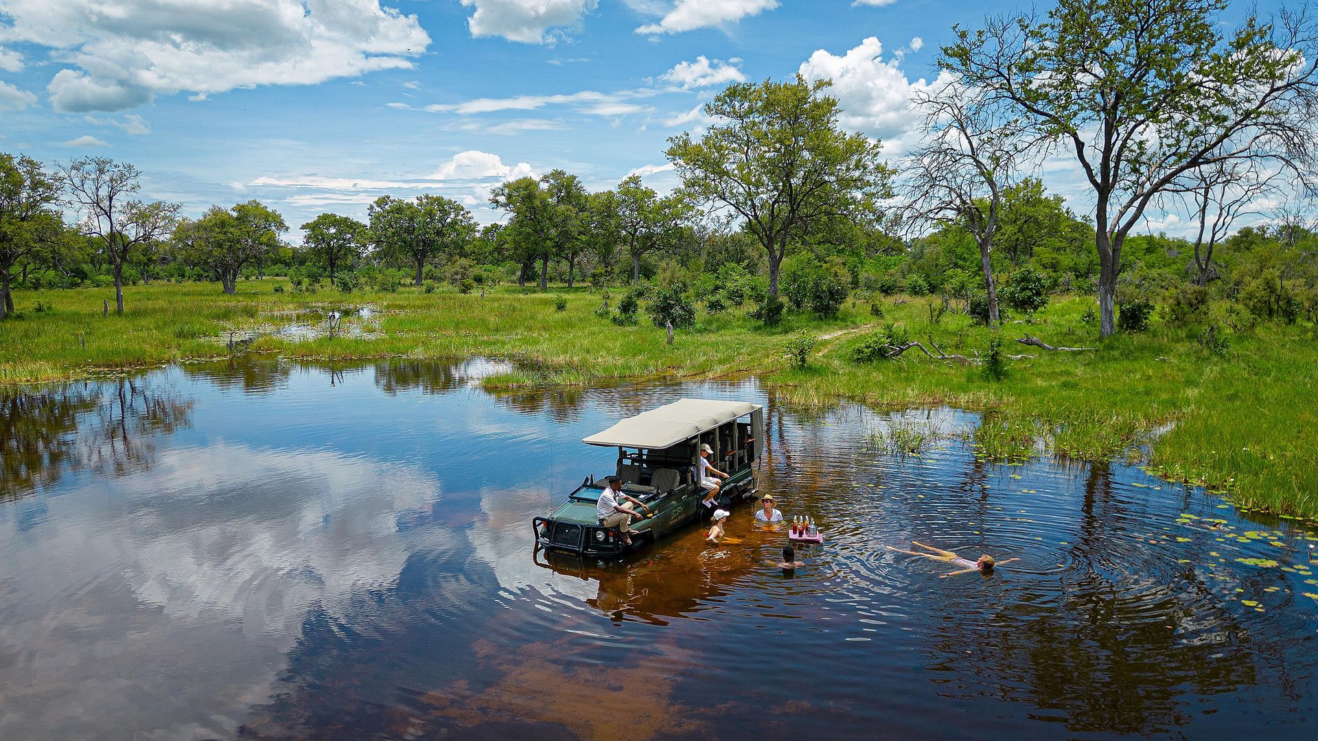 An open-topped safari vehicle drives through a shallow, reflective body of water in a lush green African savanna.