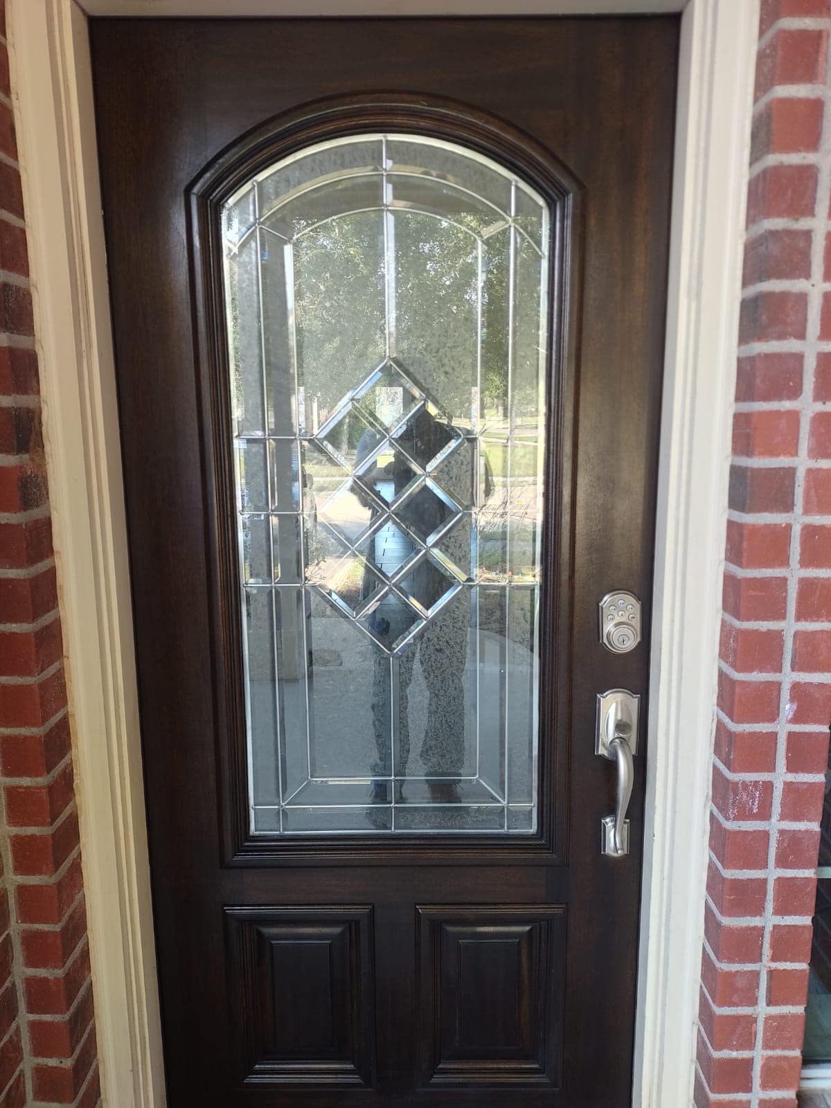 Dark wooden front door with decorative glass panel and silver handle beside a brick wall.