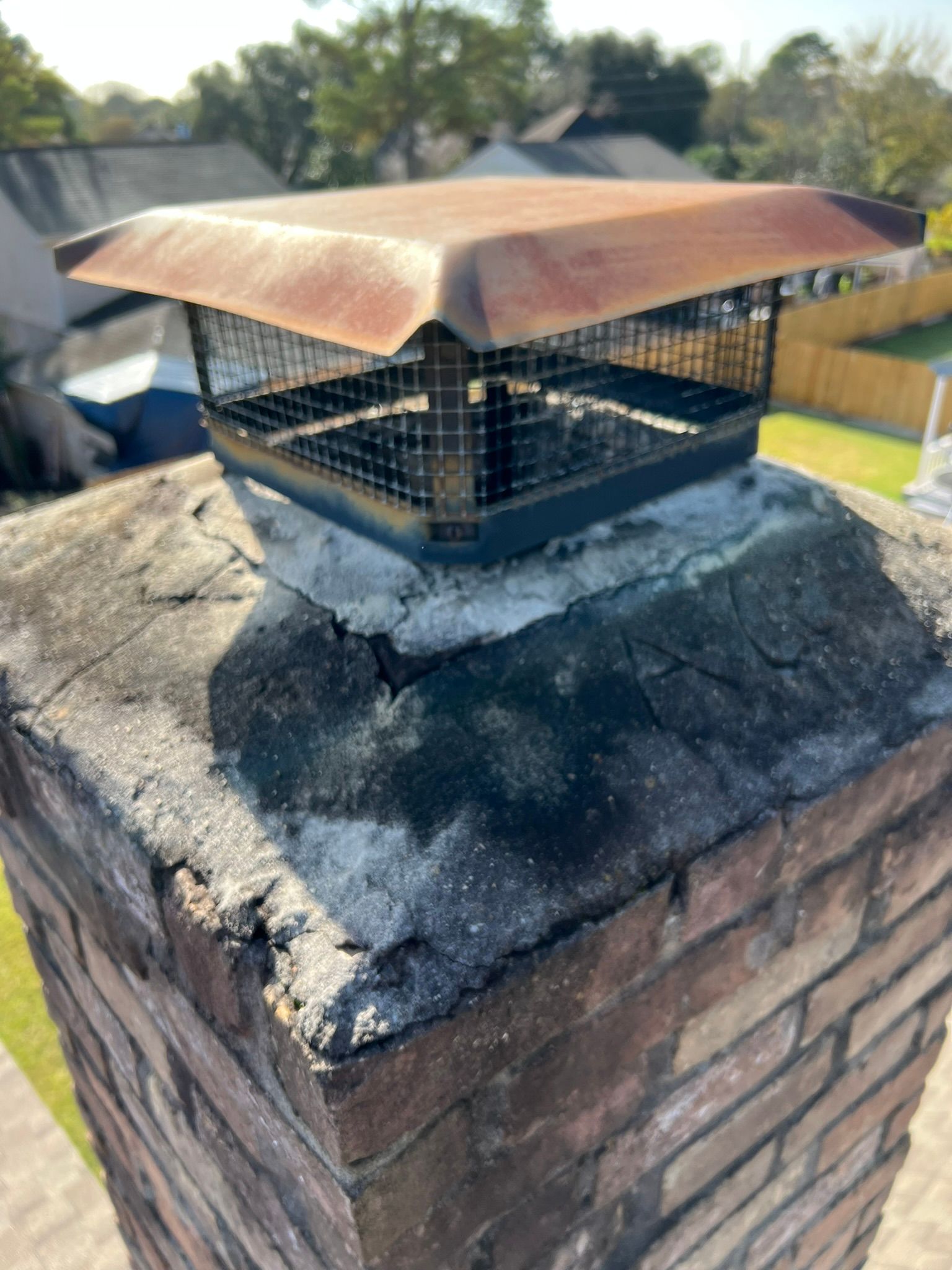 Rusted metal chimney cap on a brick chimney with soot stains and a roof in the background