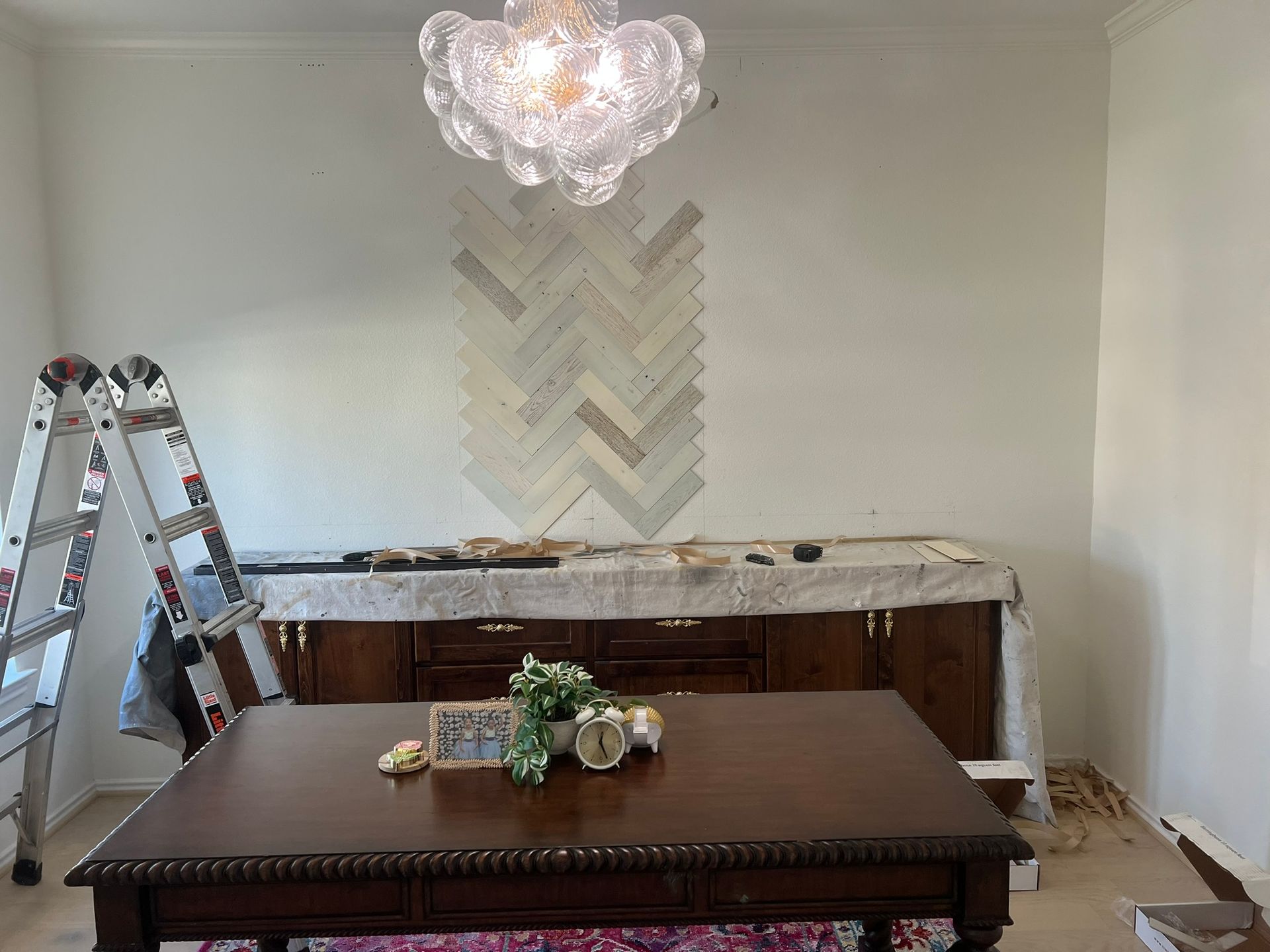 Dining room under renovation with ladder, unfinished cabinet, and table in front of herringbone wall art and chandelier