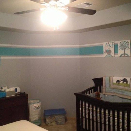 Nursery room with gray walls, teal border, crib, dresser, and ceiling fan.