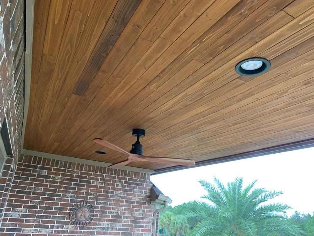 Wood-paneled porch ceiling with a black fan and recessed light above a brick wall and palm tree.