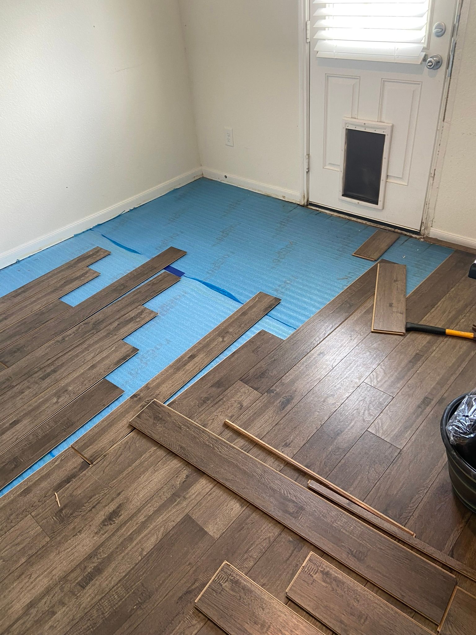Room with blue underlayment and wood flooring planks being installed near a door.