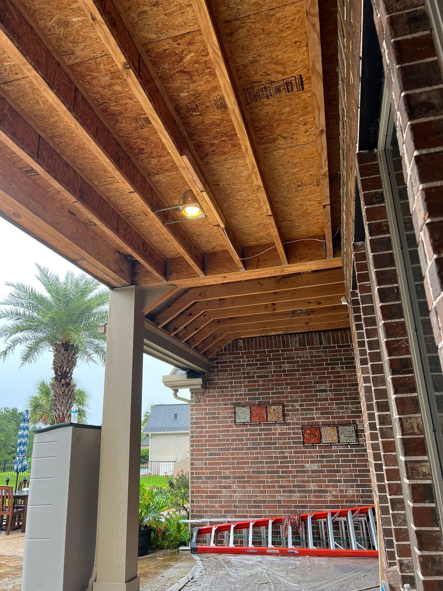Brick patio under a wooden pergola with stacked red chairs, a ladder, and a palm tree outside