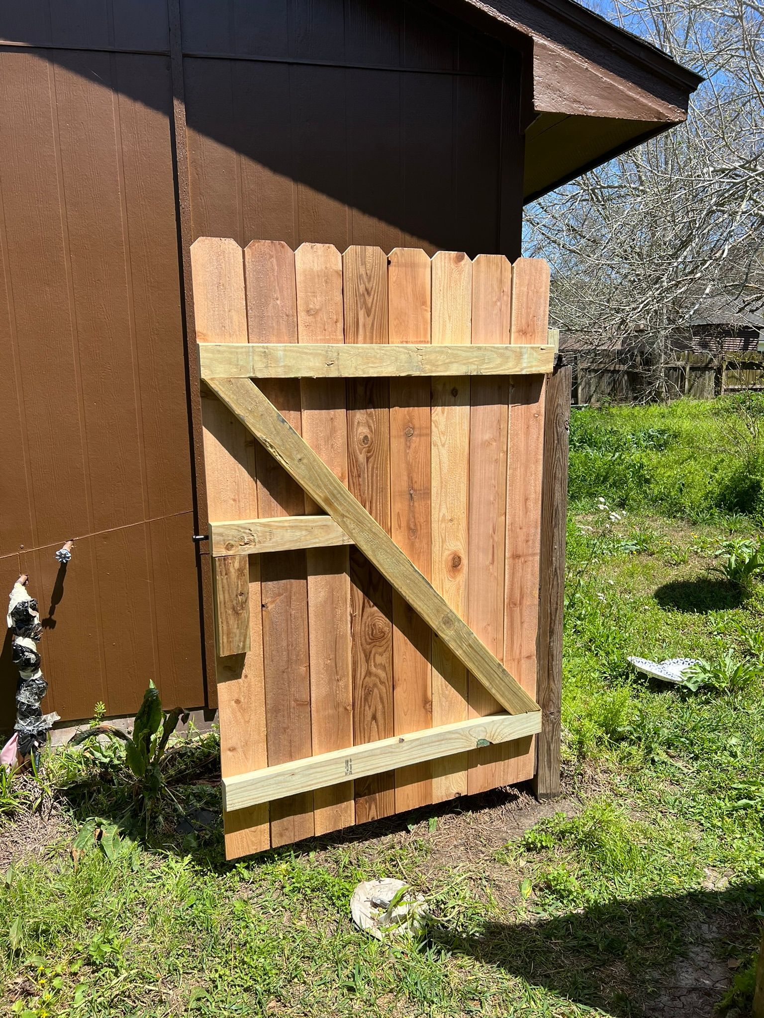 Wooden fence gate with diagonal brace beside a brown shed in a grassy yard.