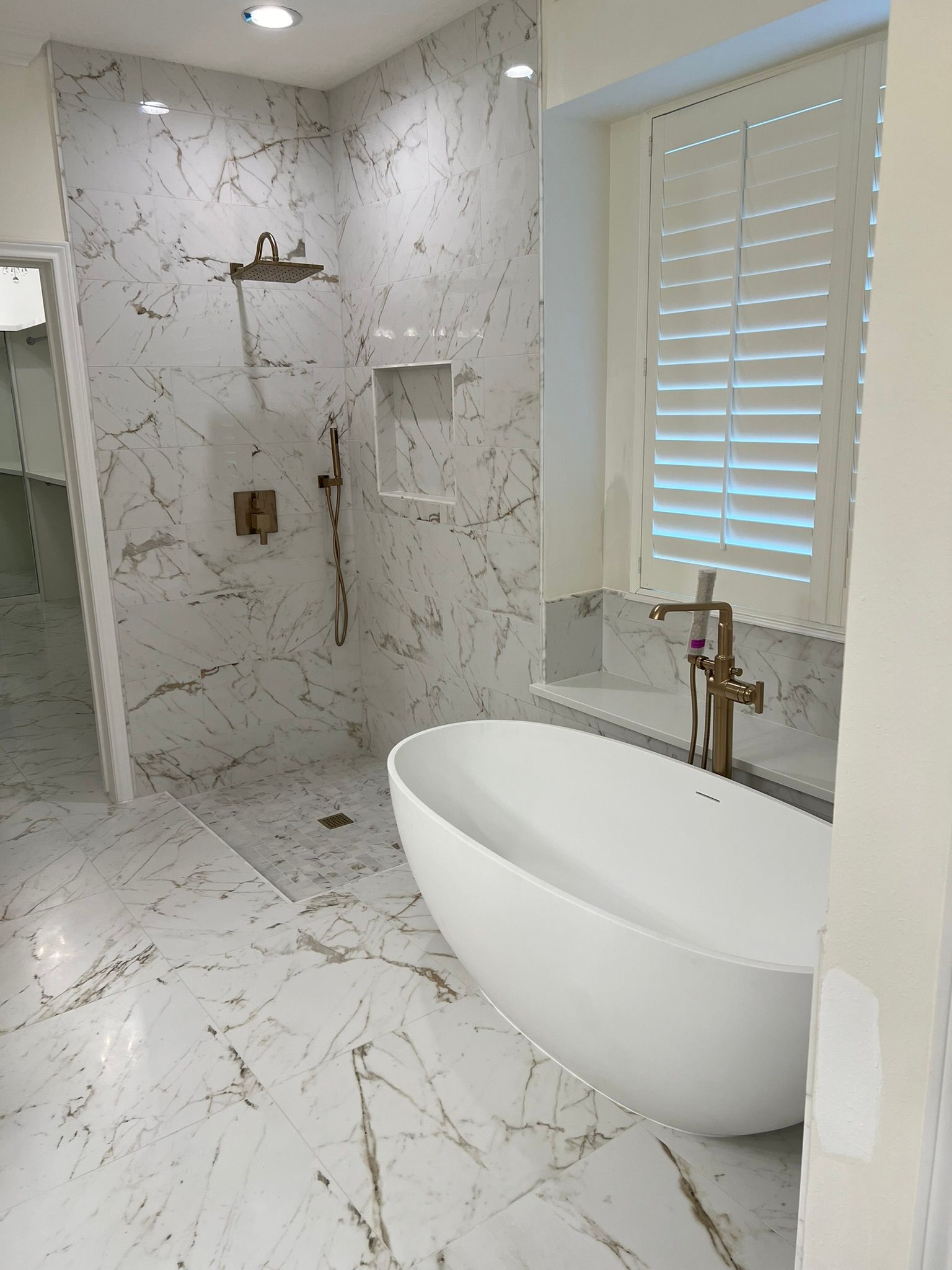 Modern white bathroom with marble tile, freestanding oval tub, and shower area near a window with blinds