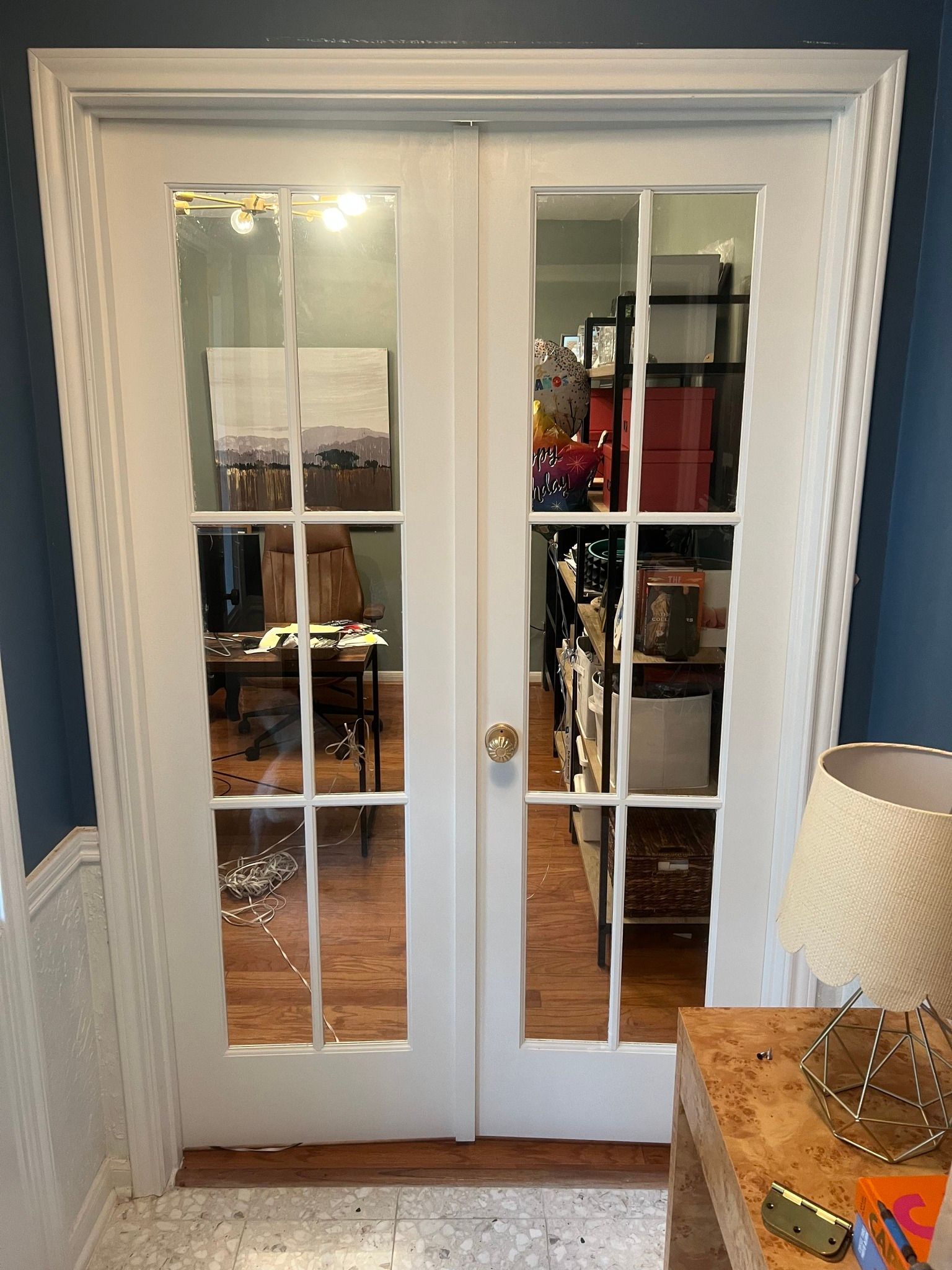 White French doors with mirror panes in a blue room, reflecting furniture and a lamp beside a wooden cabinet.
