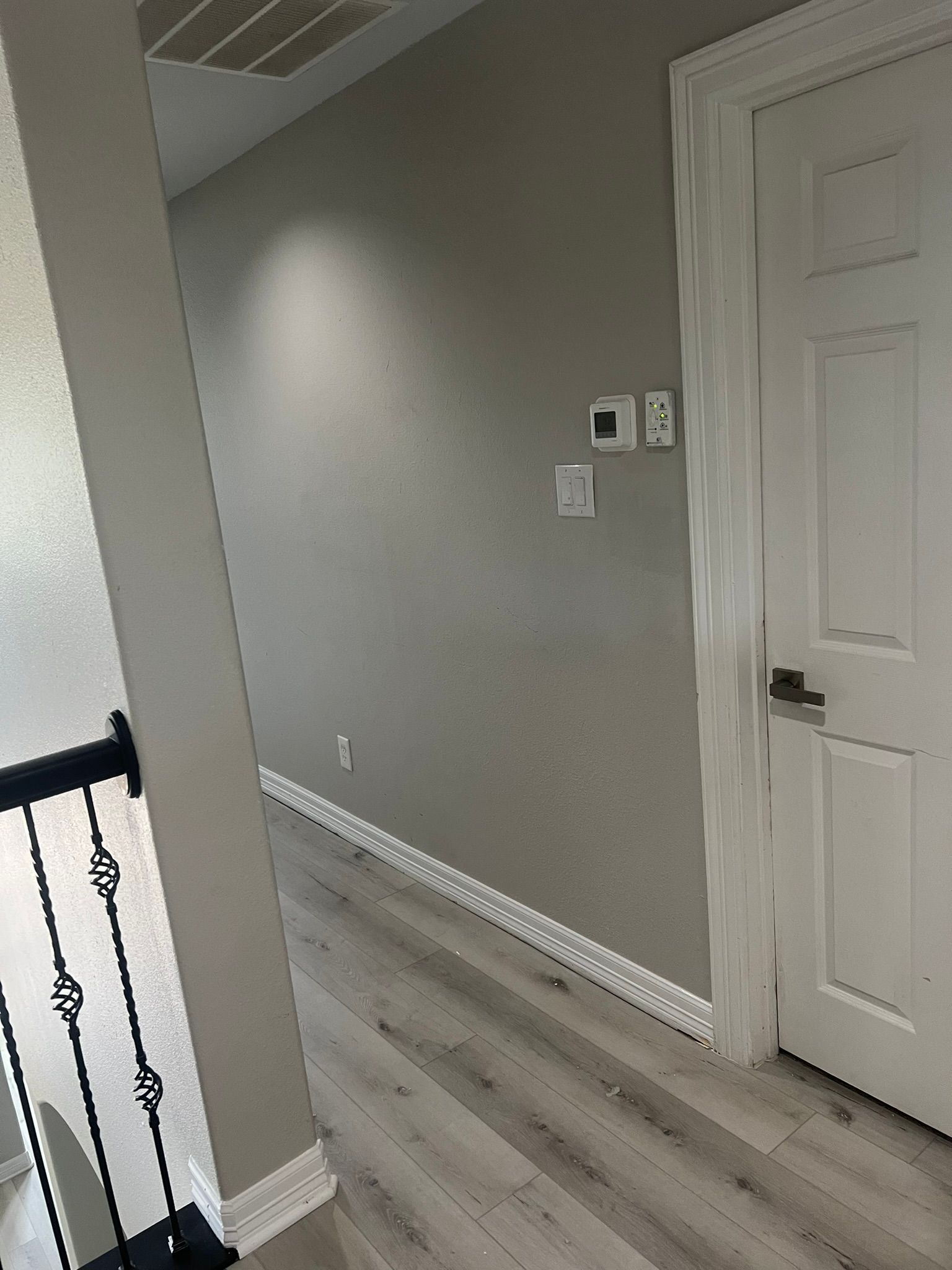 Narrow hallway with gray walls, white door, and light wood flooring; stair railing at left.