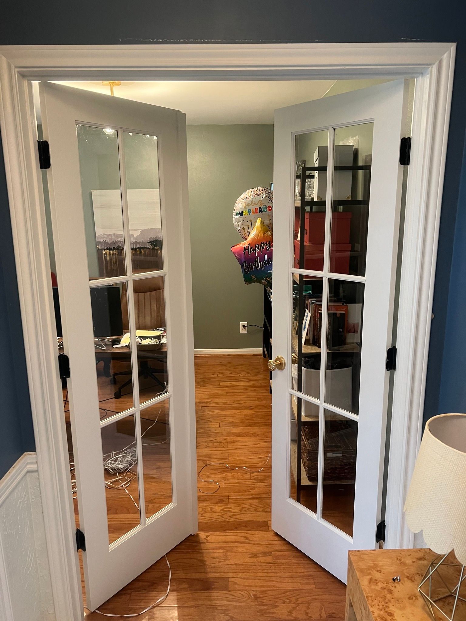 Open French doors into a room with hardwood floors, a shelf, and a colorful hanging item.