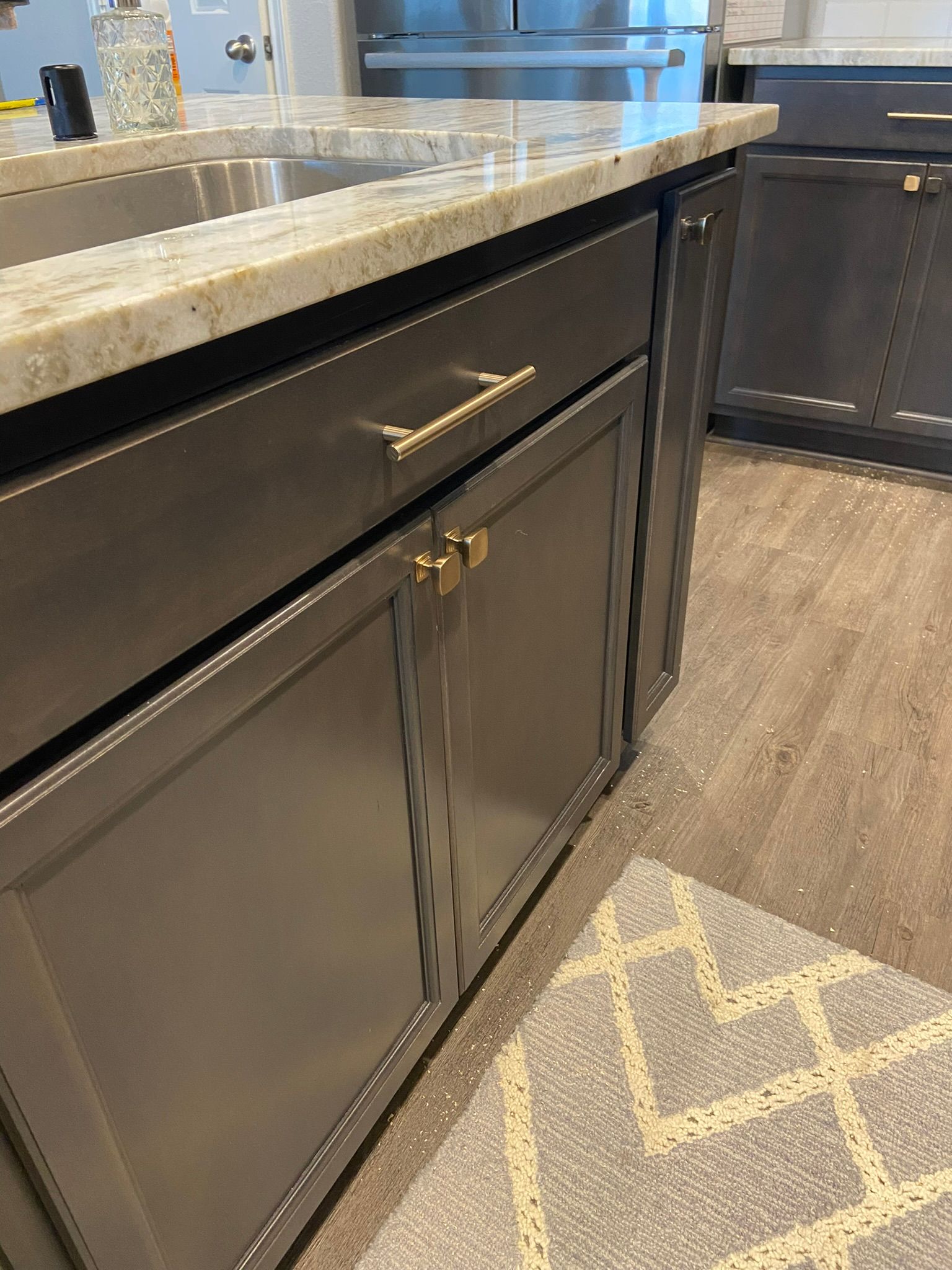 Dark kitchen cabinet with brass handles beneath a speckled countertop and patterned rug.