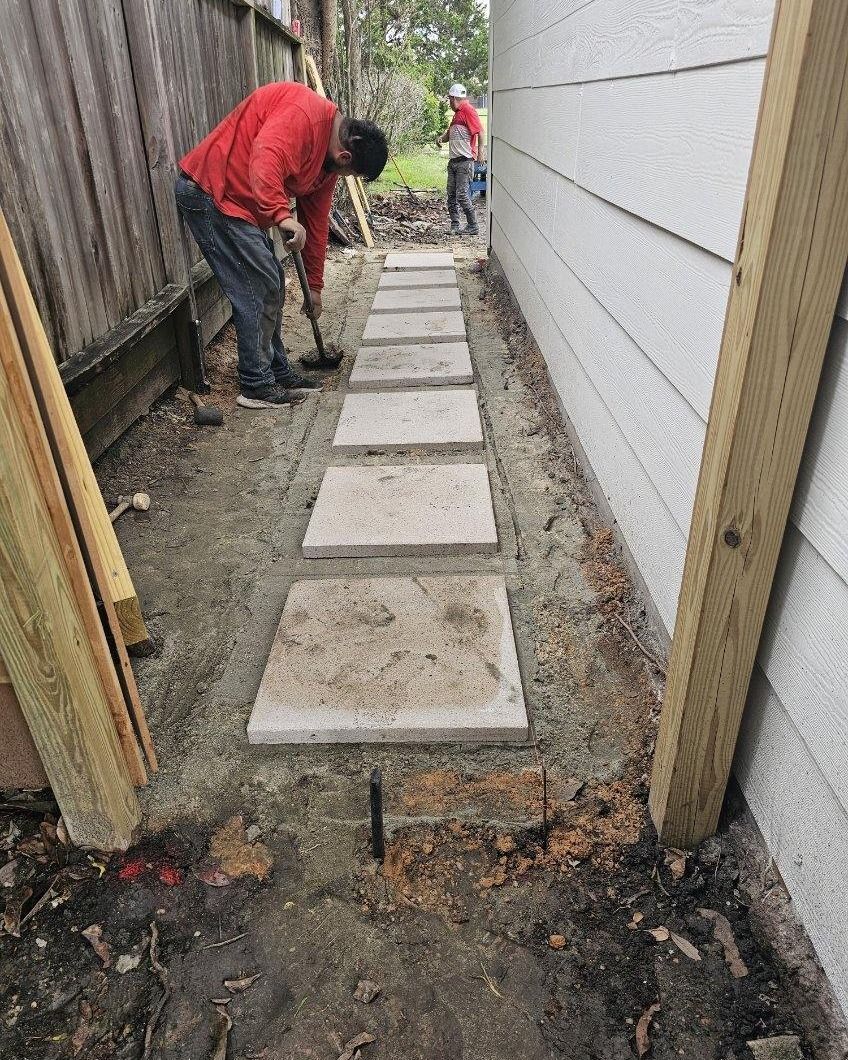 Narrow side yard with newly laid concrete stepping stones, a worker in red, and dirt borders alongside a house