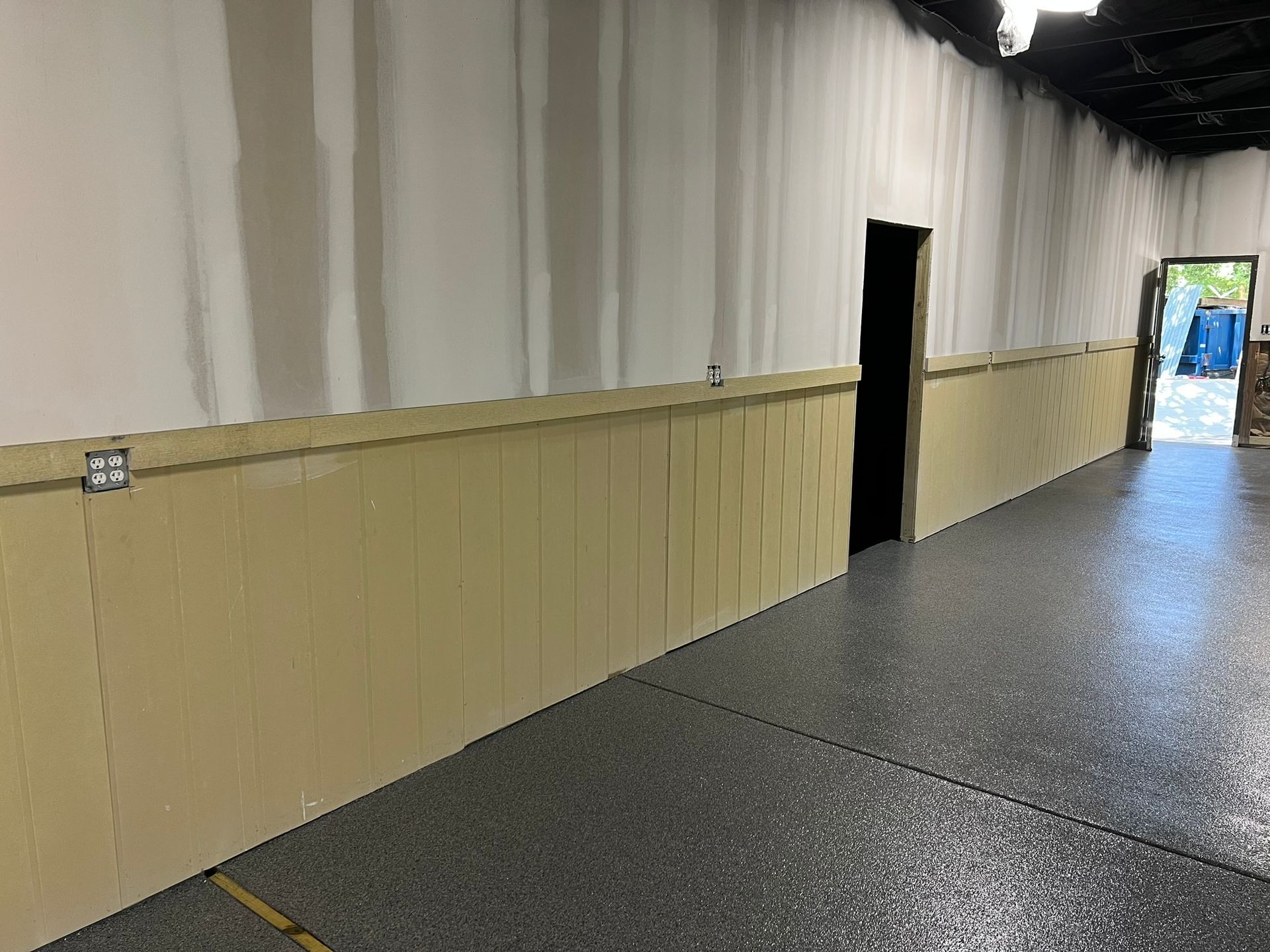 Empty indoor hallway with beige walls, dark flooring, and an open doorway to daylight at the end
