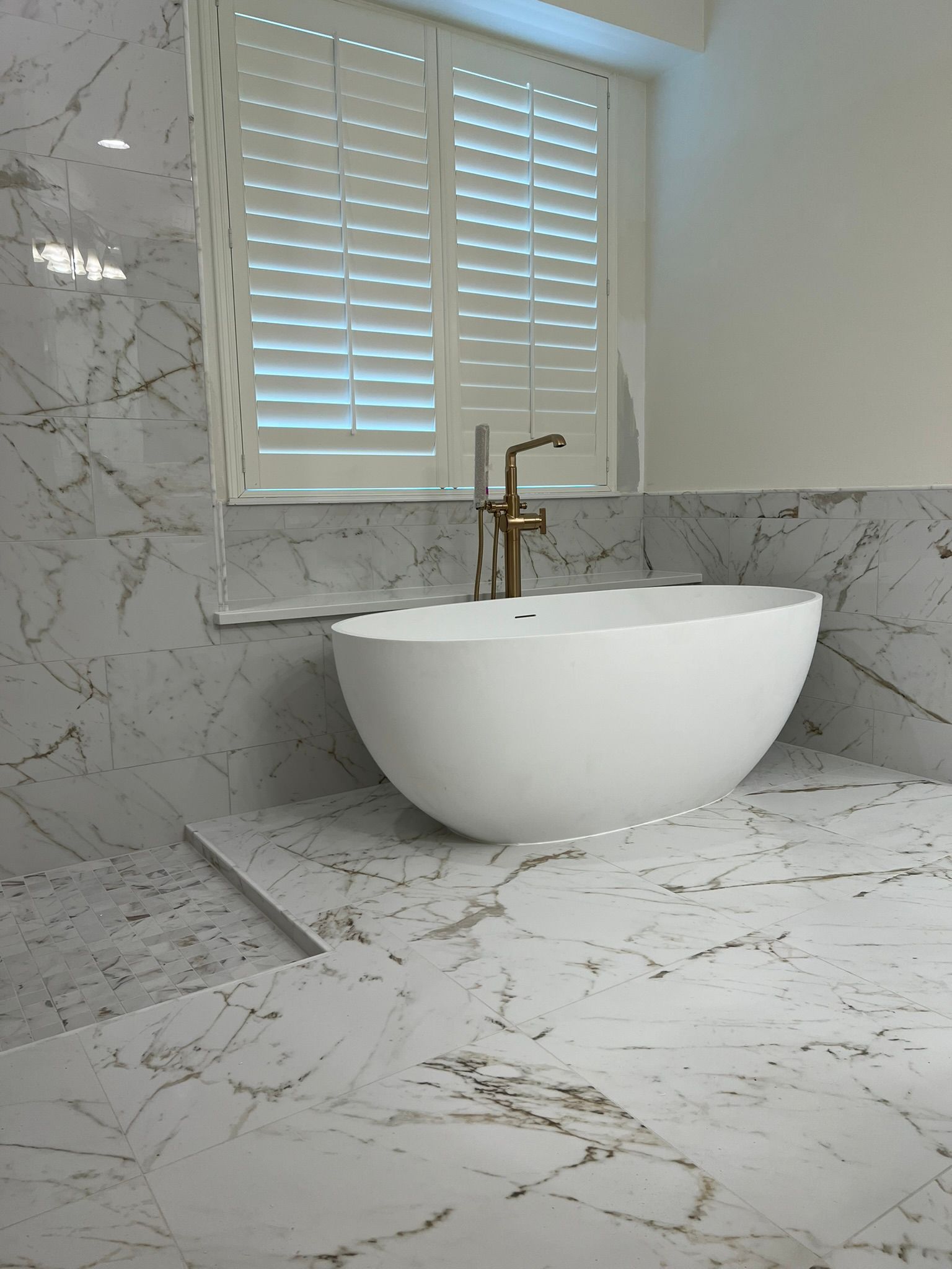 Modern bathroom vanity with white vessel sink, marble walls and countertop, and a window with shutters.