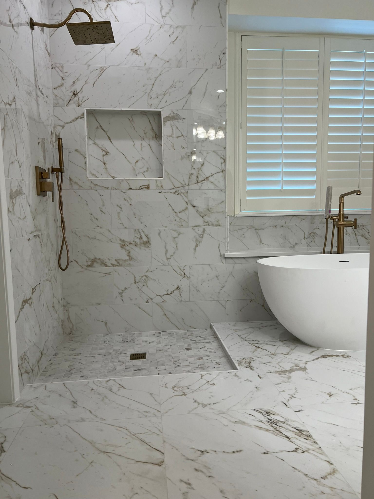 Modern white marble bathroom with walk-in shower, window shutters, and a freestanding tub