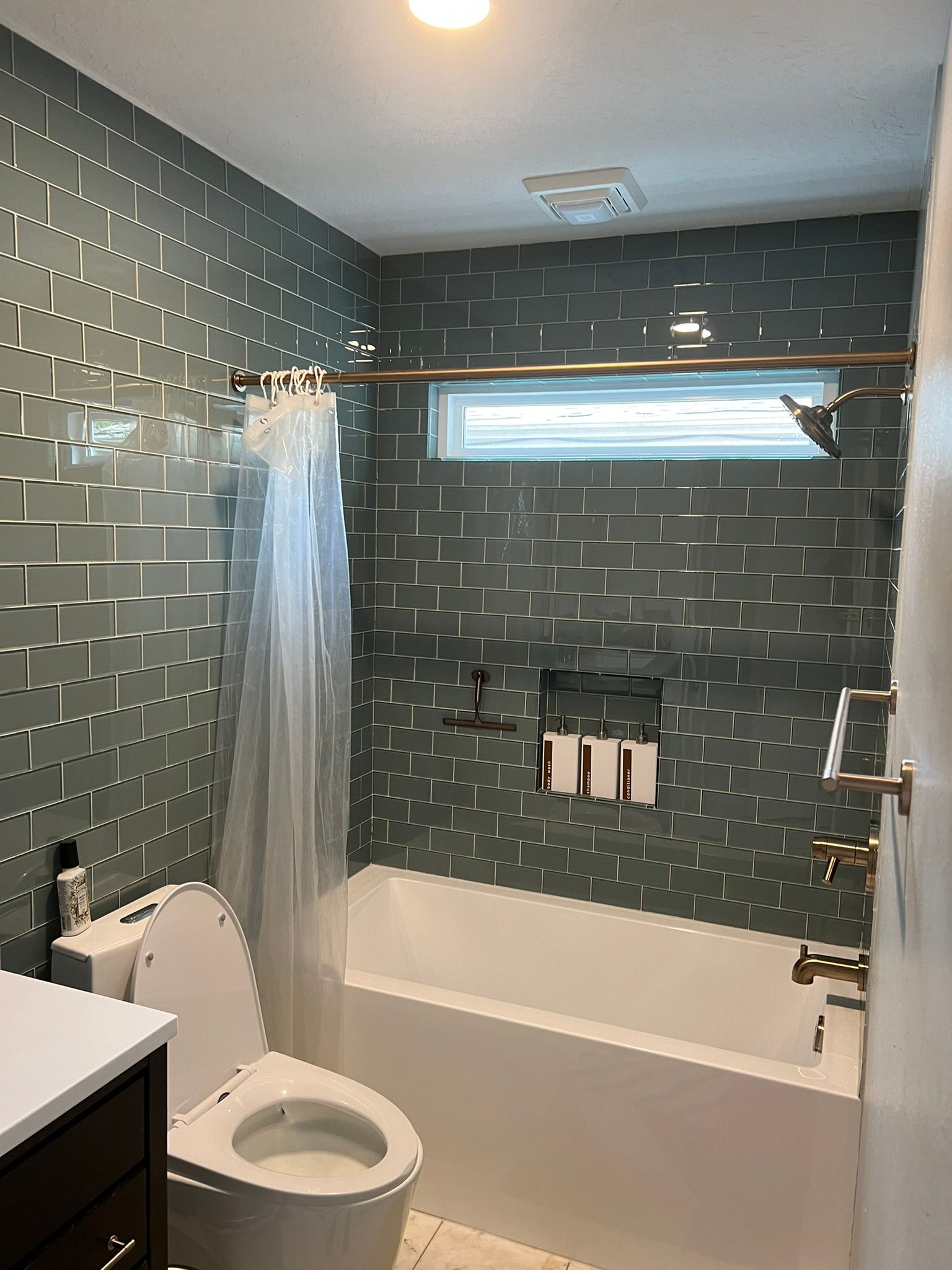 Small bathroom with green tile walls, bathtub, toilet, shower curtain, and a high window.