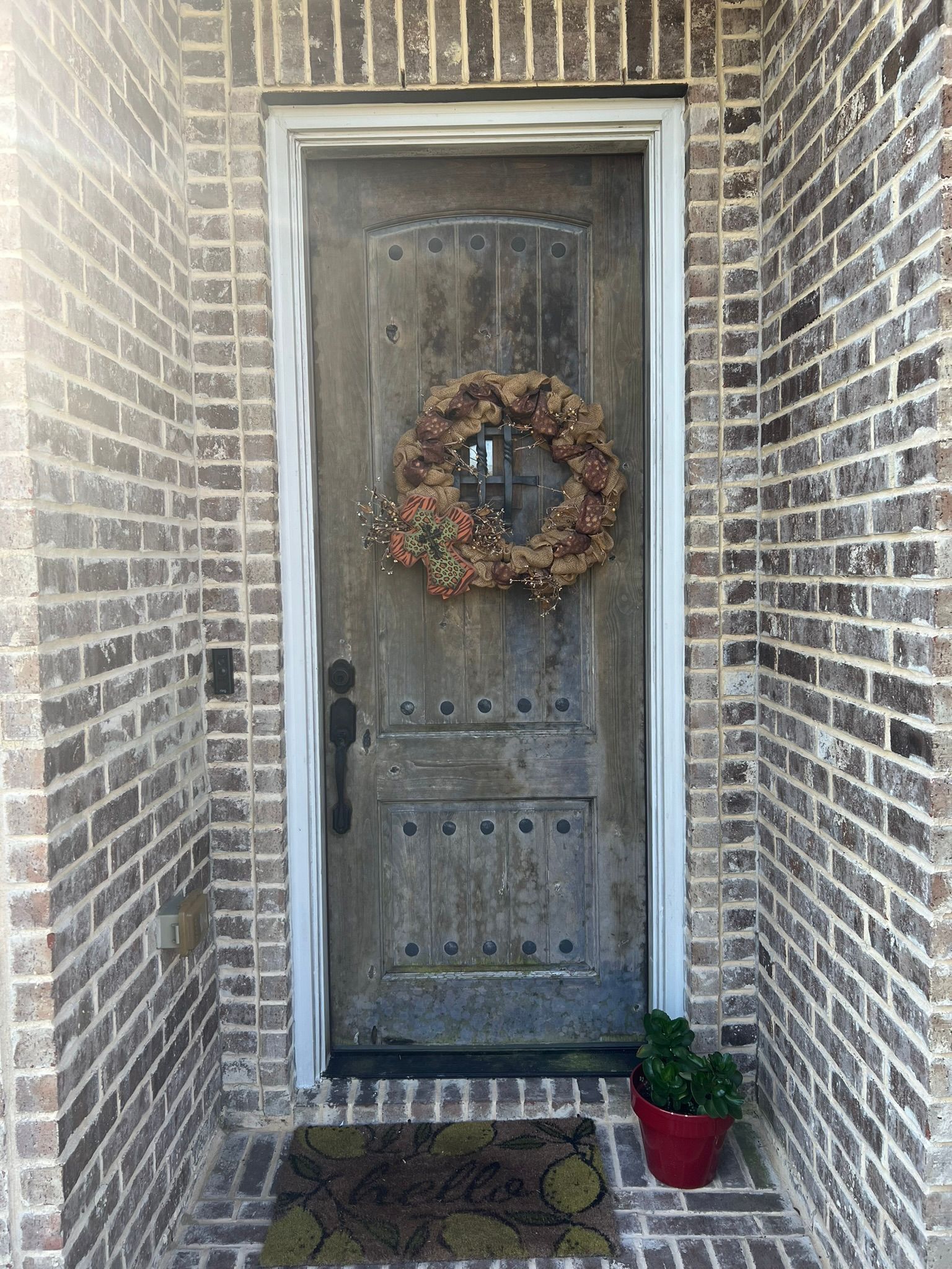 Rustic gray front door with wreath, framed by brick walls and small potted plants at the entrance