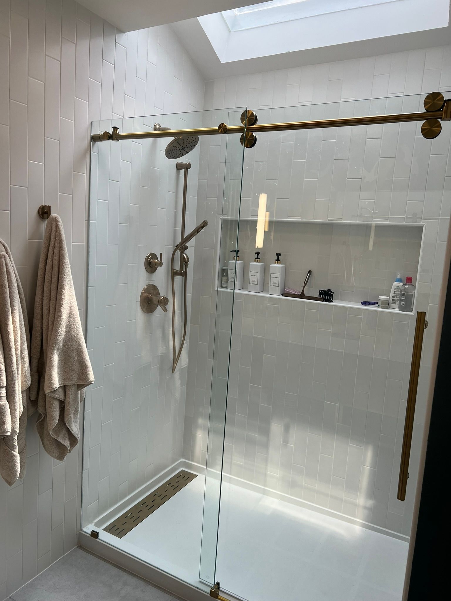 Walk-in shower with glass door, brass shower fixtures, white tile walls, and hanging towels