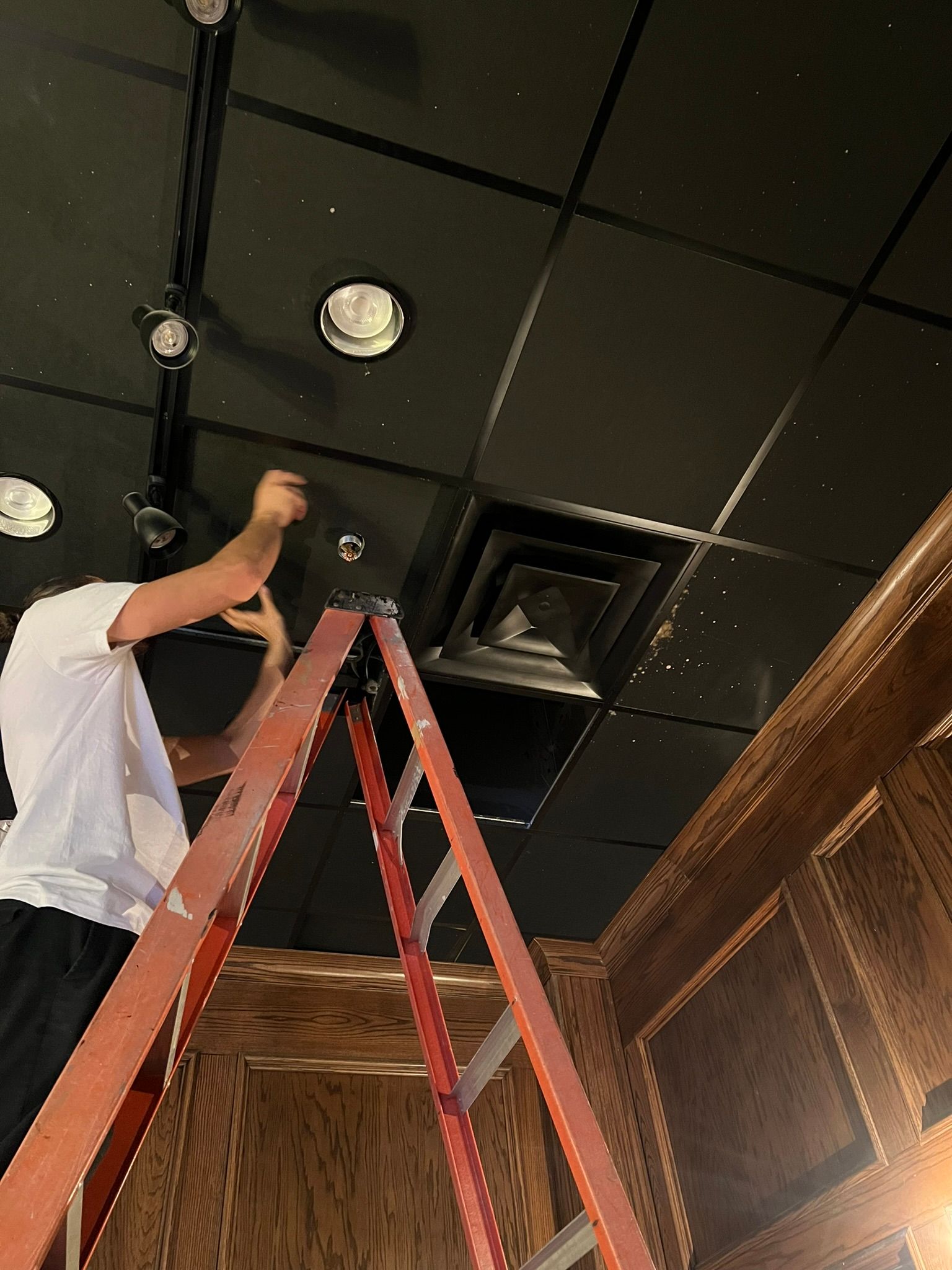 Person on a ladder repairing a black ceiling with recessed lights and vents in a wood-paneled room