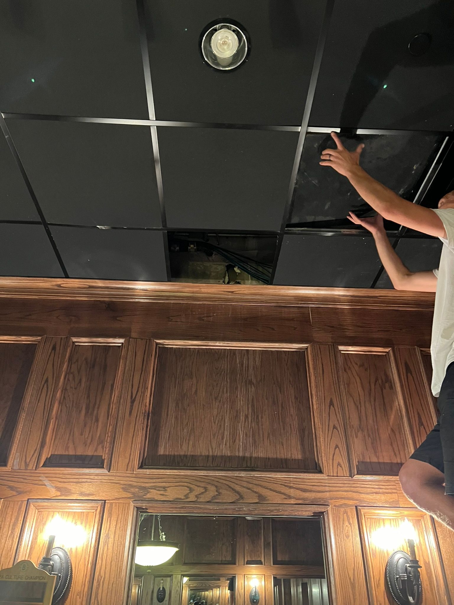 A person points upward at a dark ceiling above a wood-paneled room with wall lights.