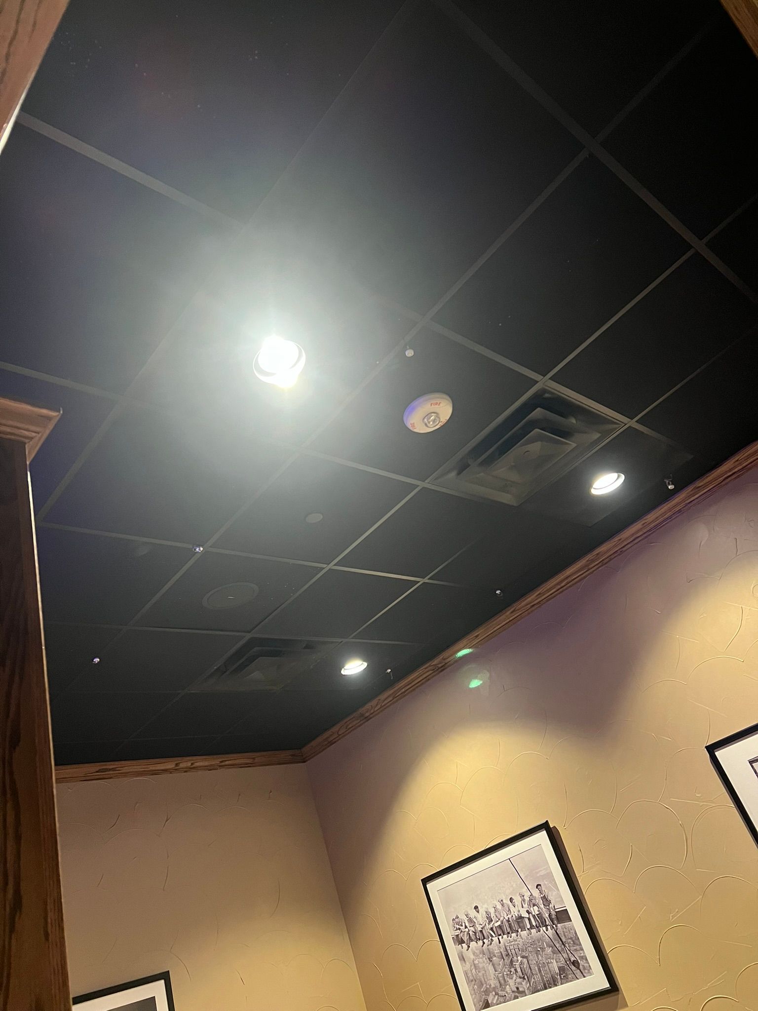 Dim room ceiling with bright lights, dark grid panels, beige walls, and framed artwork.