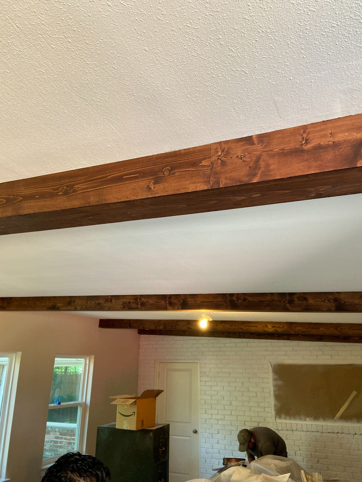 Living room with exposed wooden beams, white brick wall, and a person bending over near a fireplace.