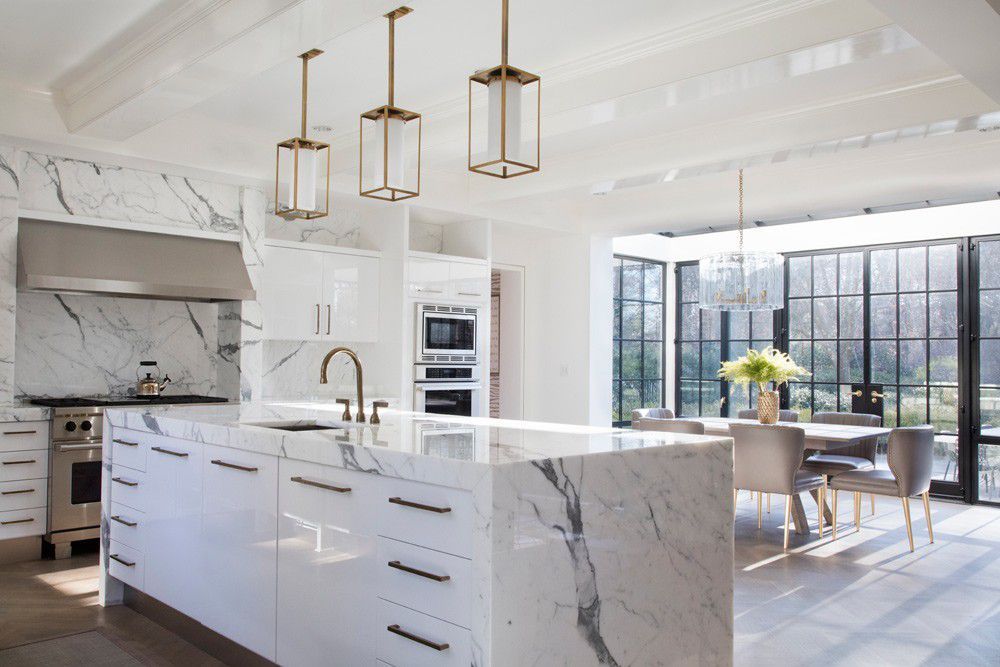 Bright modern kitchen with marble island, brass pendant lights, and large windows overlooking a dining area