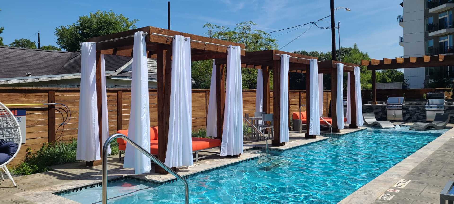 Poolside cabanas with white curtains beside a turquoise swimming pool on a sunny day
