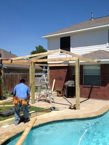 Two workers building a wooden poolside pergola beside a backyard pool and two-story house