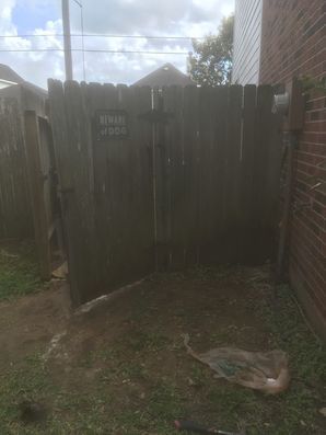 Open wooden gate in a narrow dirt yard beside a brick wall, with a rock in the foreground.