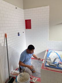 Worker installing white wall tiles in a bathroom, with tools, bucket, and covered sink in view