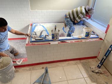 Two workers tiling a bathroom tub surround with white subway tiles and red grout lines