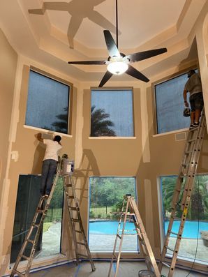 Workers on ladders painting a tall beige living room with a ceiling fan and pool view.
