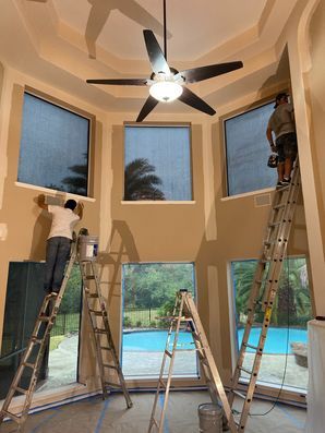 Two workers on ladders installing windows and trim in a bright room with a ceiling fan and pool view.