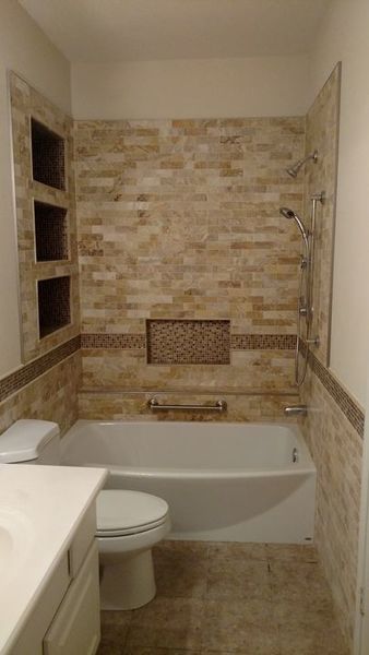 Small beige tiled bathroom with bathtub, toilet, vanity edge, and built-in wall shelves.