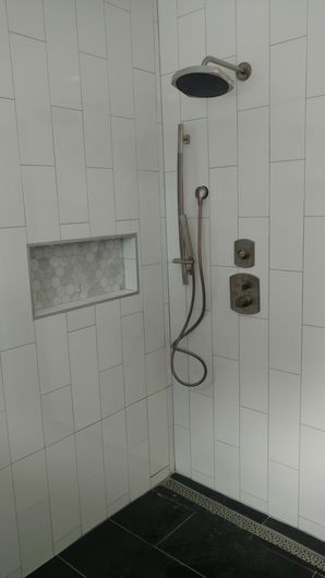 Walk-in shower with white tile, black floor, built-in shelf, and handheld showerhead and controls