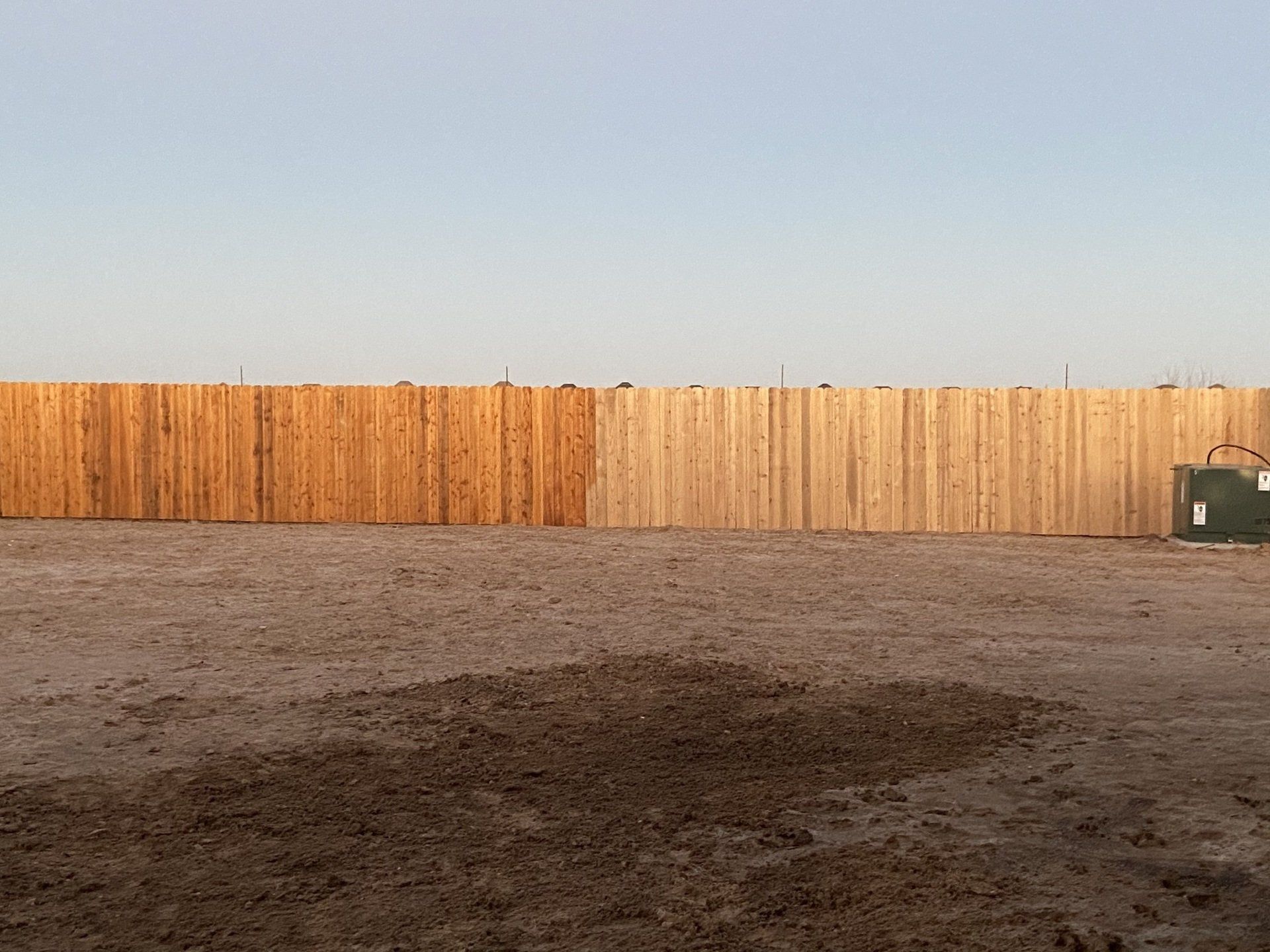 Empty dirt lot with a tall wooden fence and a dumpster under a clear sky