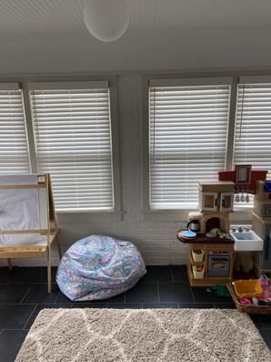 Playroom corner with bean bag, toy kitchen, crib, and windows with blinds over a rug and dark floor