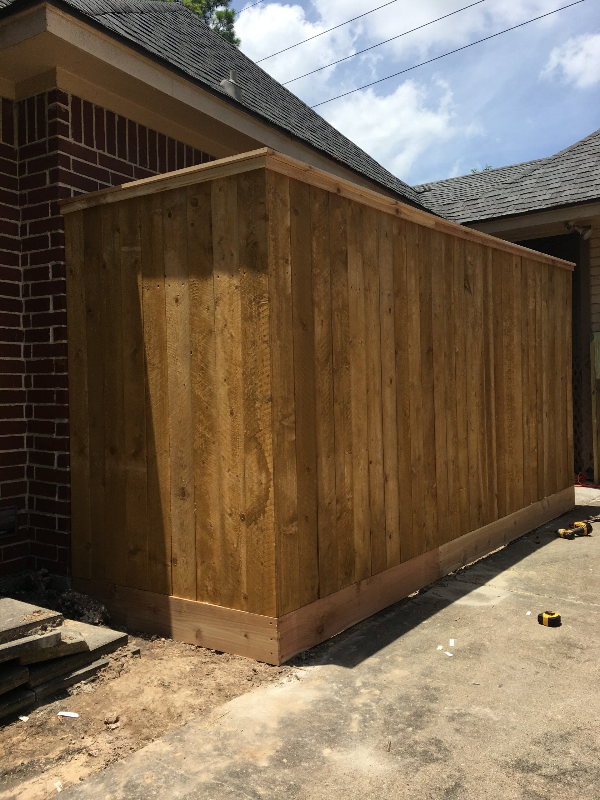 Wooden privacy fence beside a brick house and driveway under a cloudy sky