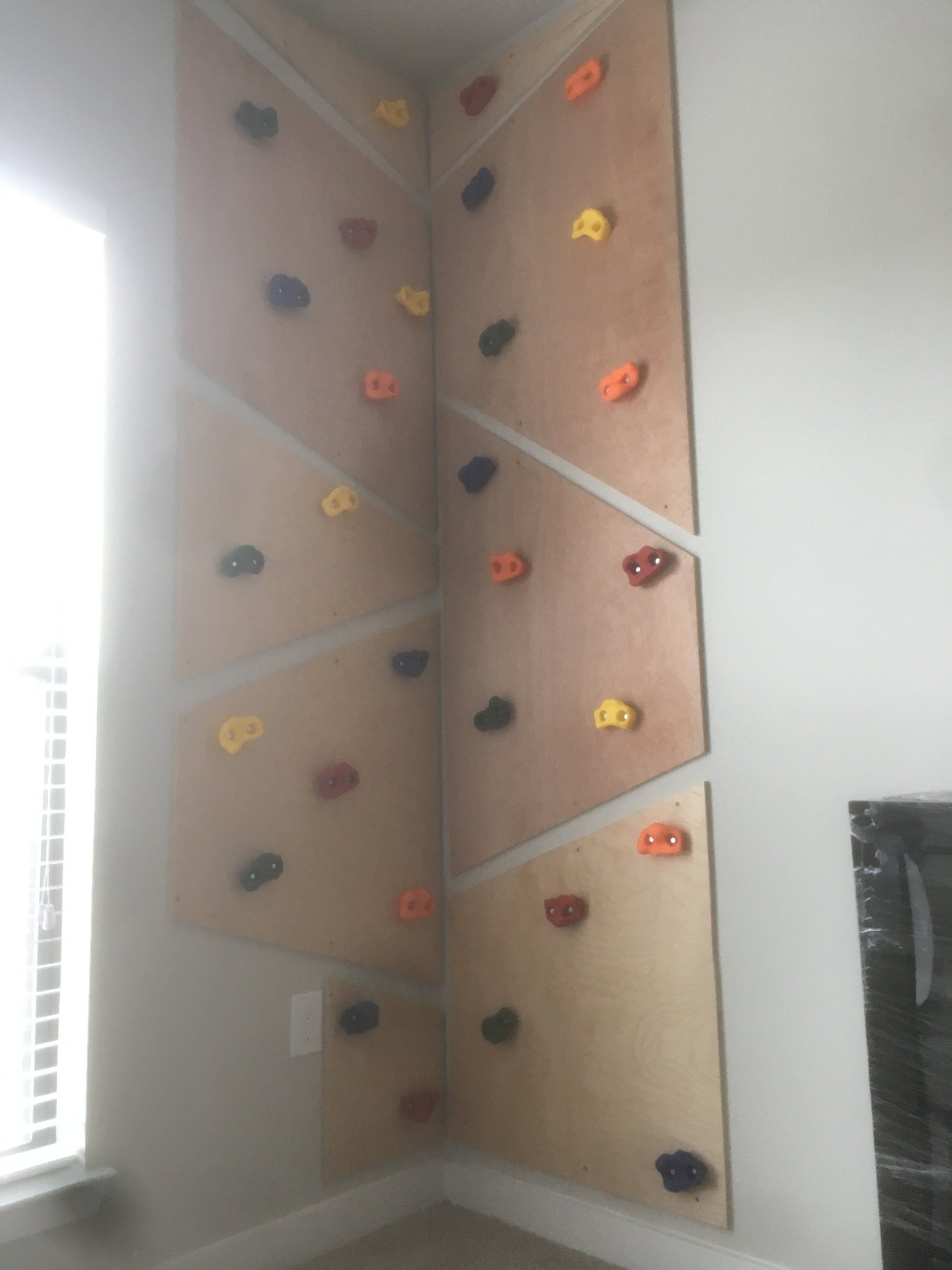 Indoor corner with beige climbing wall panels and colorful climbing holds near a bright window.