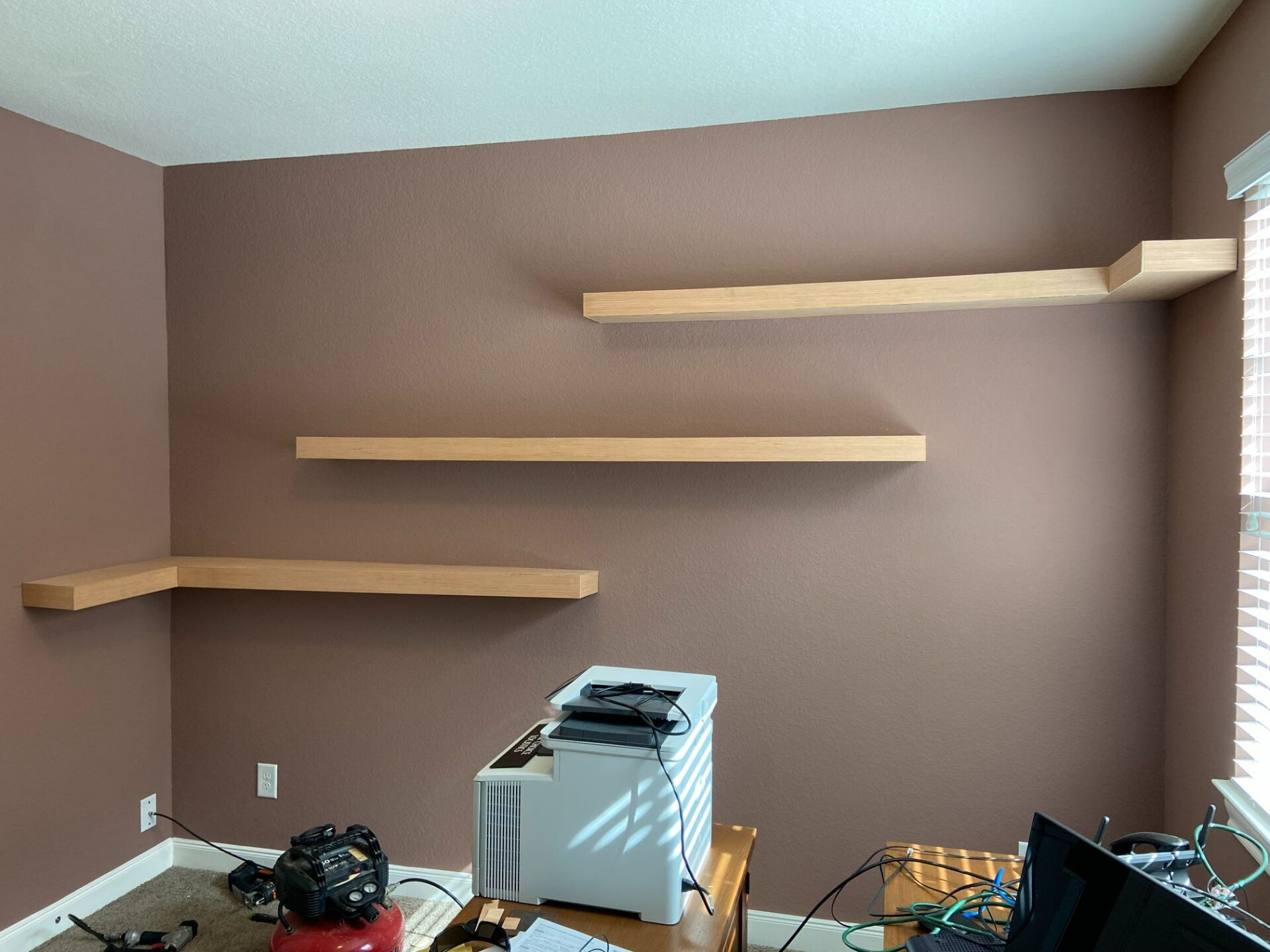Empty room with brown walls, three floating shelves, and a small printer on a desk by the window.