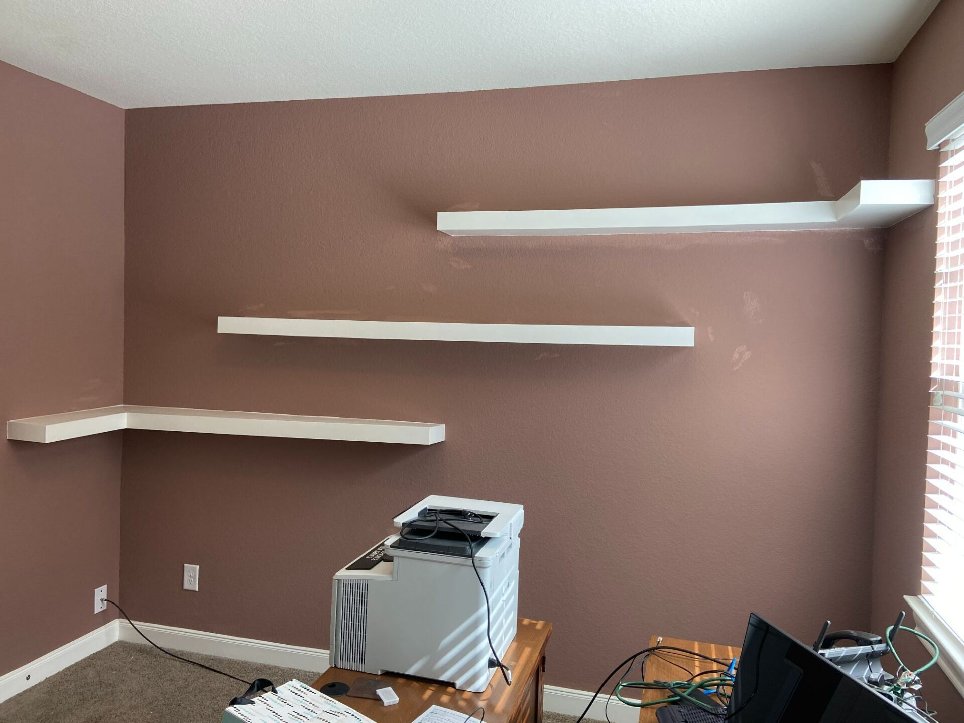 Empty room with brown walls and three white floating shelves, plus a small cabinet near the window.